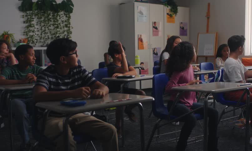 A classroom setting where several students are seated with several students raising their hands while a teacher is in the background