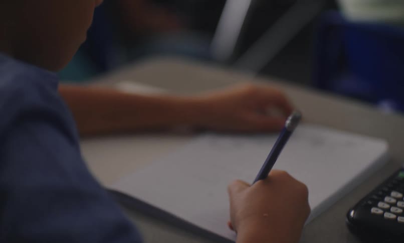 A child sitting on a desk at school writing math exercises on a notebook