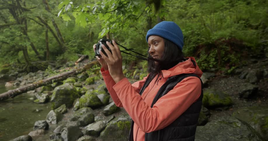 A woman outdoors wearing a blue beanie and an orange jacket photographing a scene with a camera next to a creek