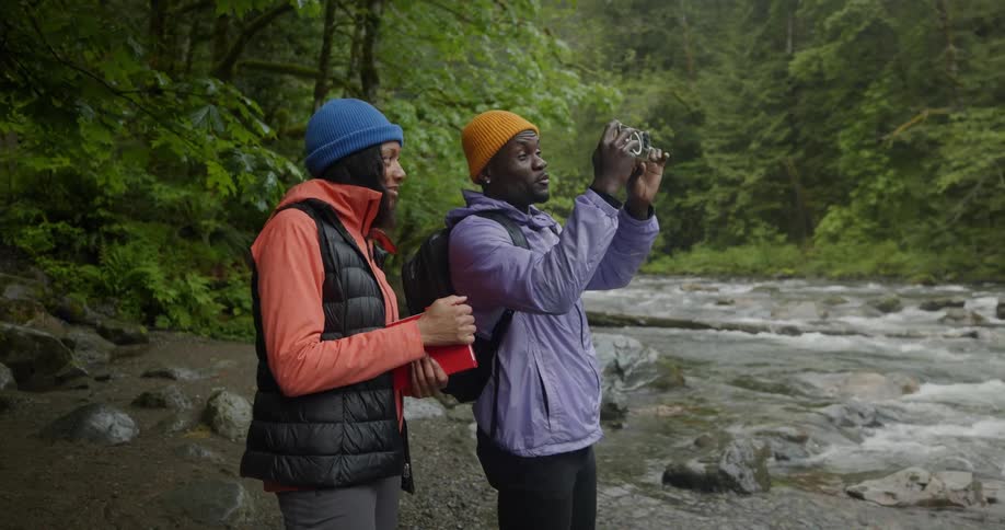 Man filming with an iphone next to a woman both talking while standing by a river in a forest