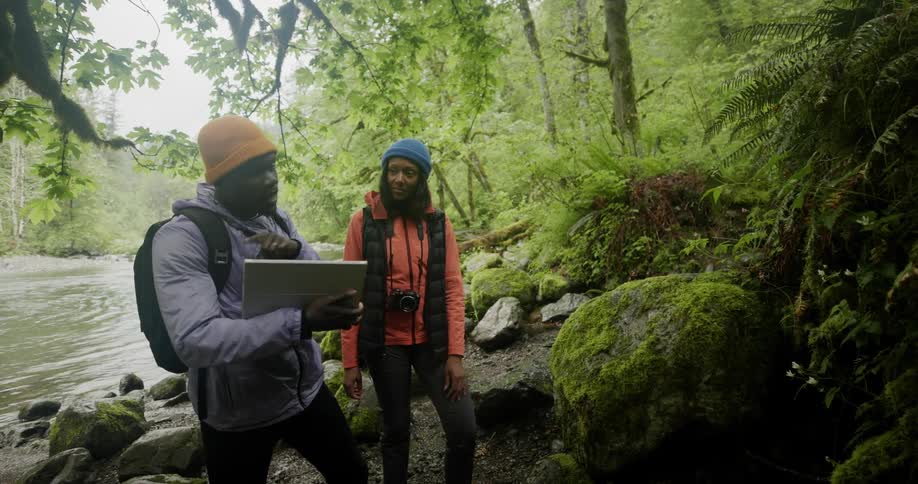 Two individuals stand near a river in a lush green forest one of them talking with a laptop in his hand