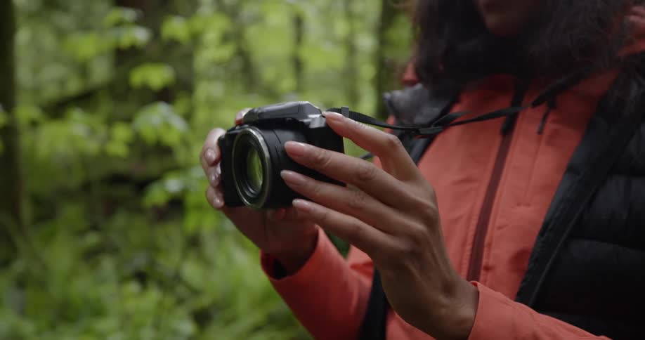 A person in a red jacket holds a black camera while standing in a forest