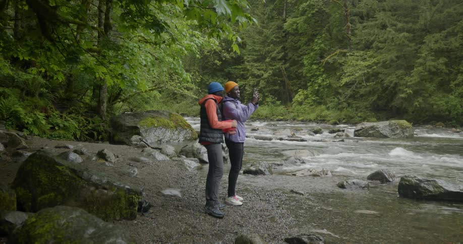 A couple stand by a river in a lush green setting taking pictures with a phone