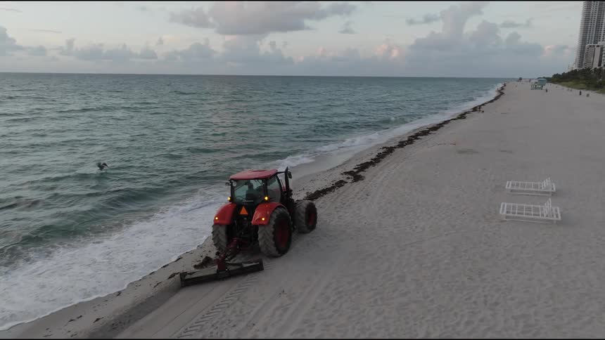 A red tractor equipped with a beach cleaning attachment drives along the sandy beach