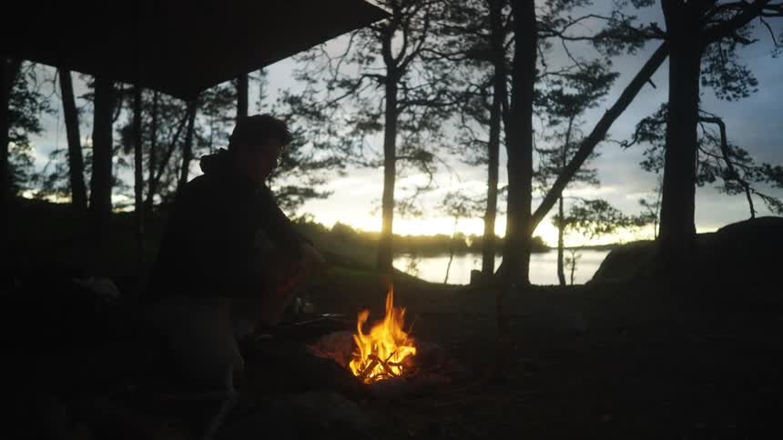 A young man builds a fire at dusk while camping above a lake