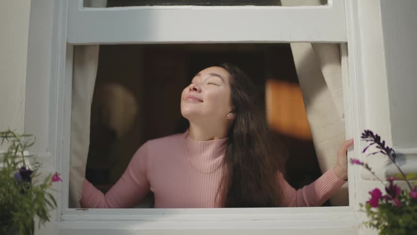 A young woman opens the curtain on her window and smells the outdoor air