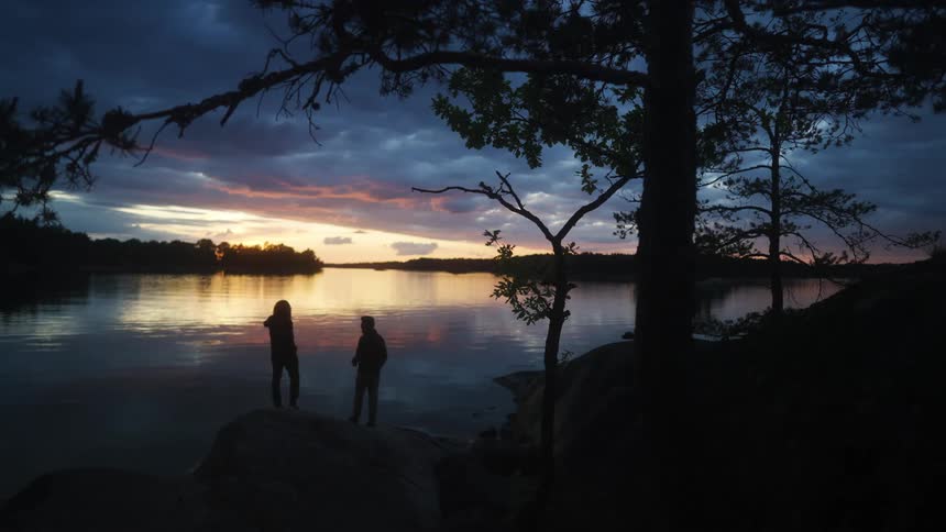 Two silhouetted figures standing by a lake at sunset surrounded by trees and a colorful sky
