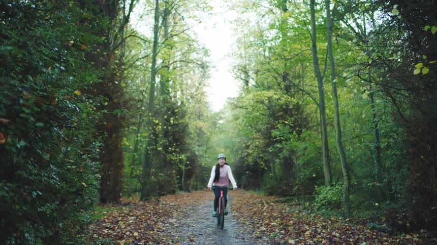 A woman wearing a helmet is riding a bicycle on a concrete path in the middle of a forest and with many dry leaves on the path