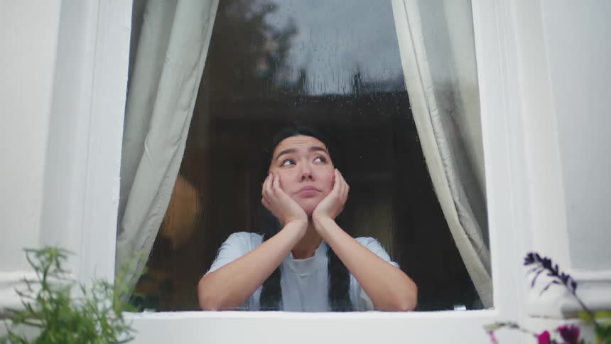 A woman lost in thought stands behind a glass window with the head on her hands