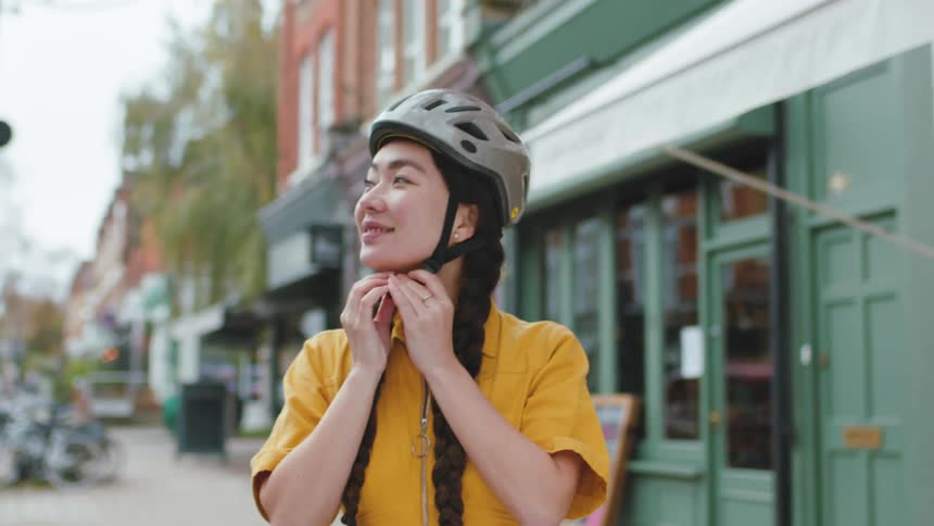 A woman in a yellow top put on a bicycle helmet standing on a city street