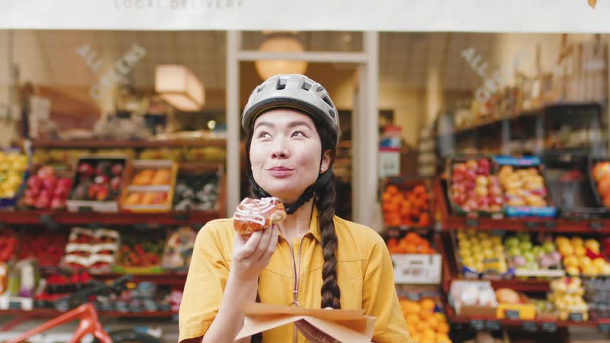 A woman wearing a bicycle helmet enjoying a pastry while standing in front of a colorful fruit and vegetable market