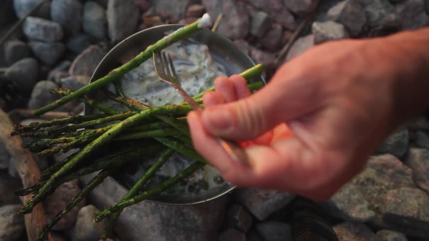 A hand picks up freshly cooked asparagus stalks from a small metal pot in a campsite