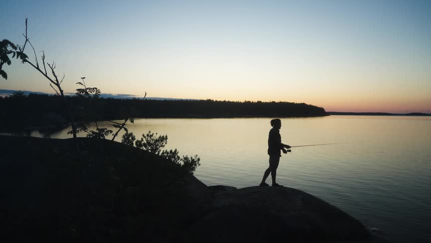 Silhouette of a person casting a fishing rod into a calm lake with a red and blue sky at dawn