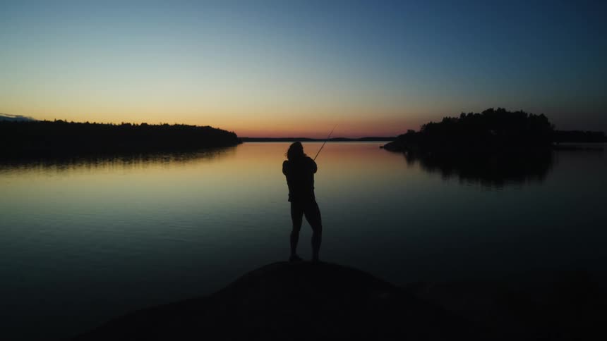 A silhouette stands at the waters edge during sunset casting a fishing line over a lake reflecting the colors of the sky