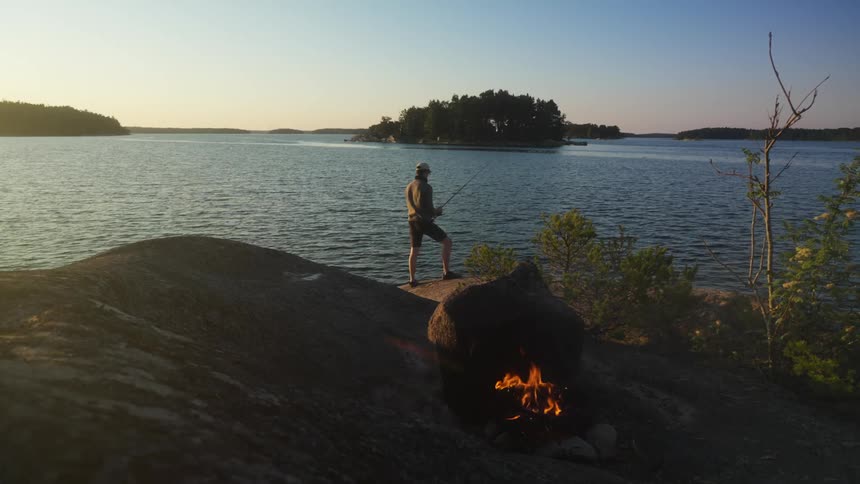 A man stands on a rocky shore near a small campfire fishing in a serene lake