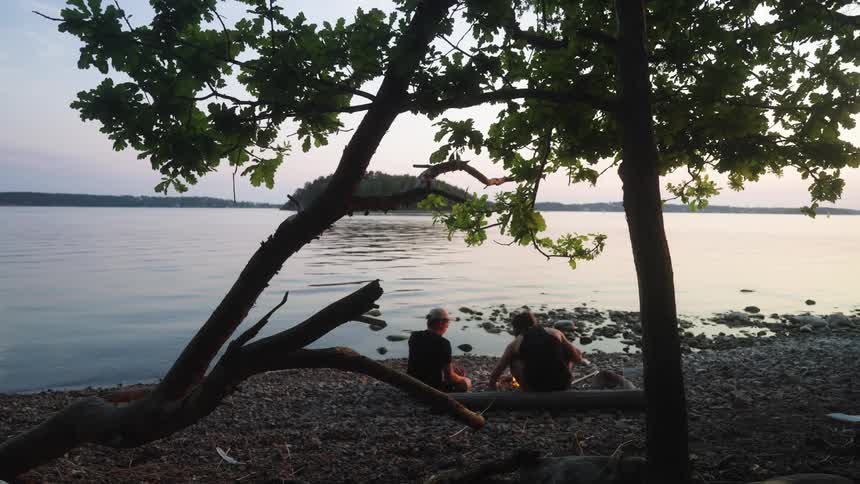 Two people sit by a lakeside a small campfire between them as dusk settles