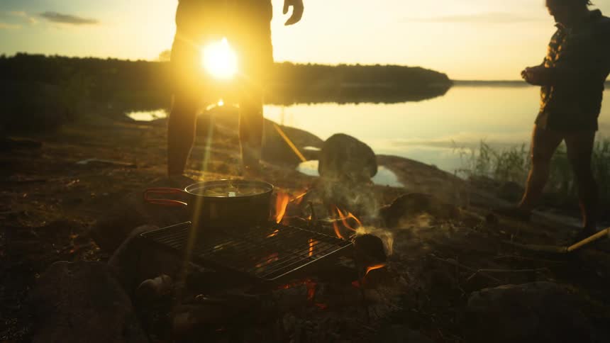 A campfire with a grill over it a pot on the grill and two people standing nearby next to a lake
