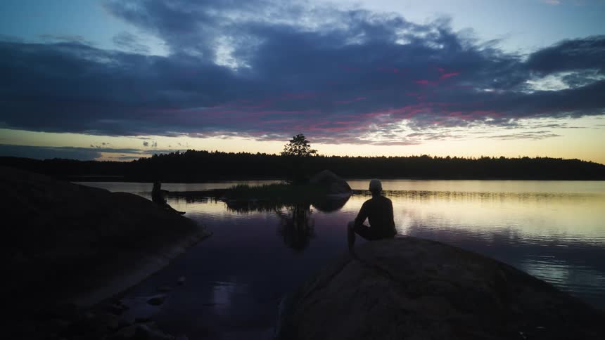 A silhouette stands on a rock by the edge of a calm lake during twilight