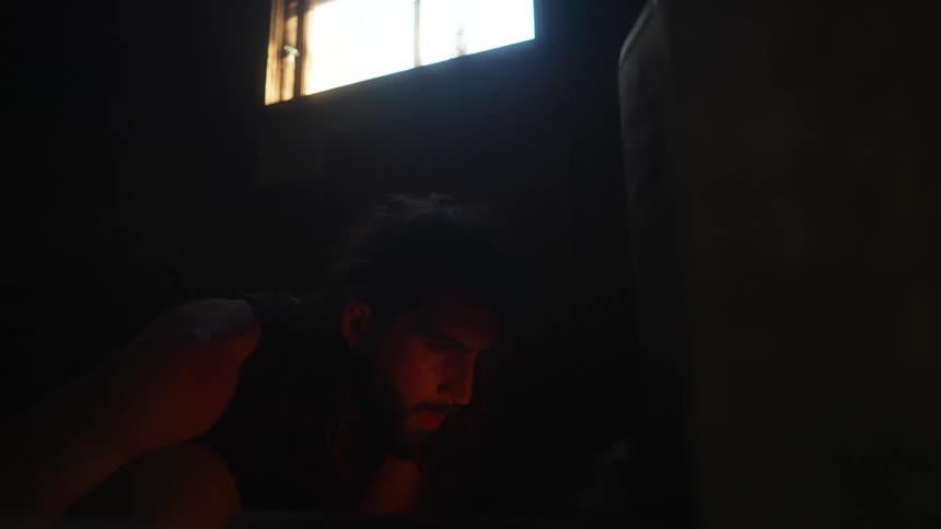 Firelight plays over the face of a man crouching in shadowed room