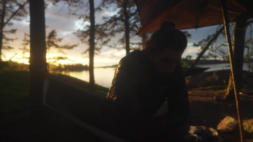 The silhouette of a man who is fixing a hammock stuck in a tree on a dark morning with the sun strong in the sky