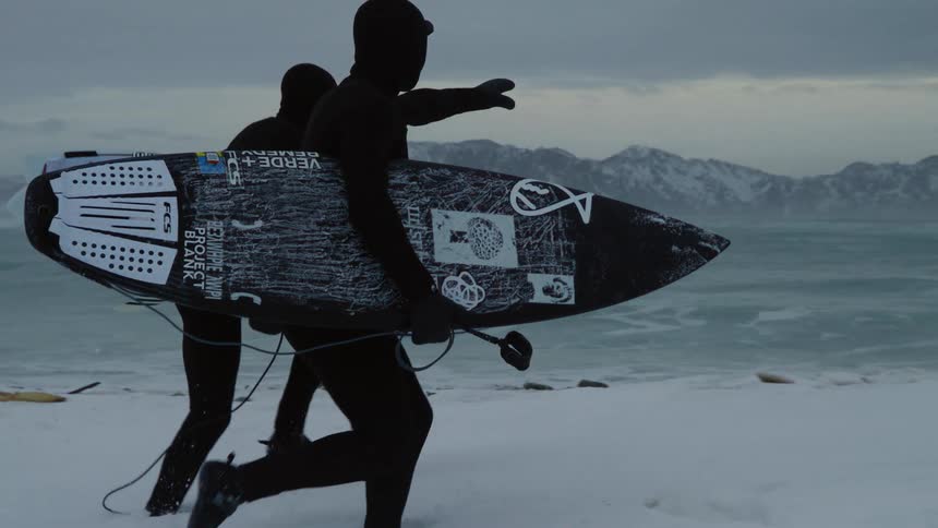 Two surfers in black wetsuits walking on a snowy beach holding surfboards with a stormy ocean and mountains in the background