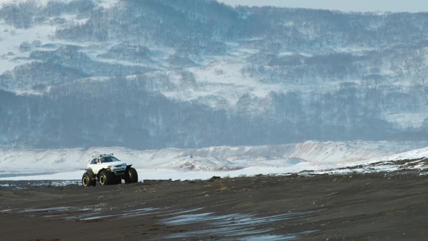 White car with giant wheels traveling on black ground with mountain ranges covered in snow in the background
