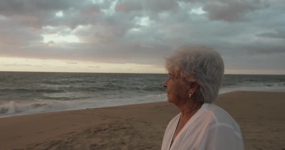 An elderly woman stands on the beach while looking out at the ocean