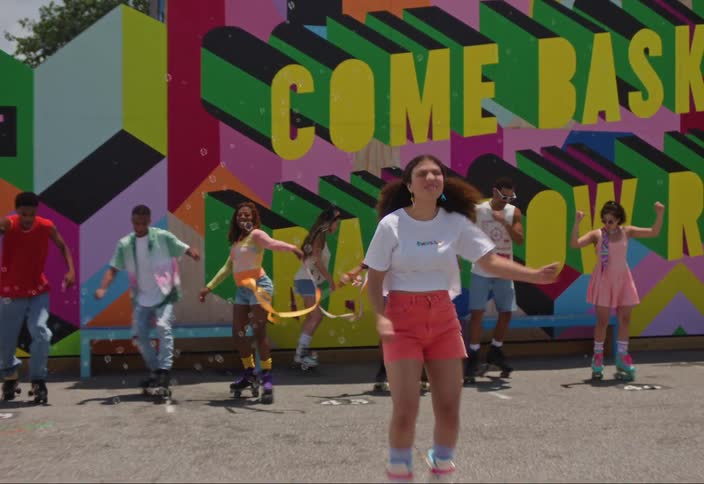 A group of people are dancing and rollerblading in front of a colorful wall in the sun