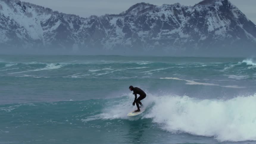 A person surfs on the waves with snowy mountains in the background