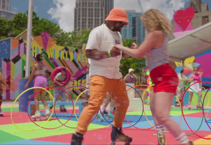 Two people are dancing together on roller skates in an outdoor roller skate park filled with colorful patterns and hula hoops