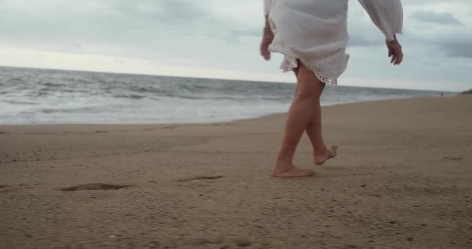 An elderly woman wearing a white blouse walks barefoot on the sand next to the beach