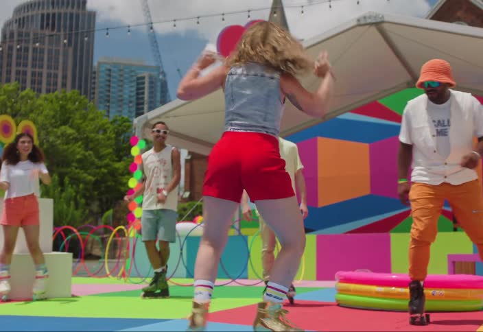 A group of friends dance while roller skating outdoors on a colorful rink with a vibrant mural backdrop