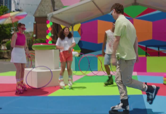 Some people in a circle and a man goes to the center and starts dancing and he is wearing rollers on his feet and the floor and background are colorful