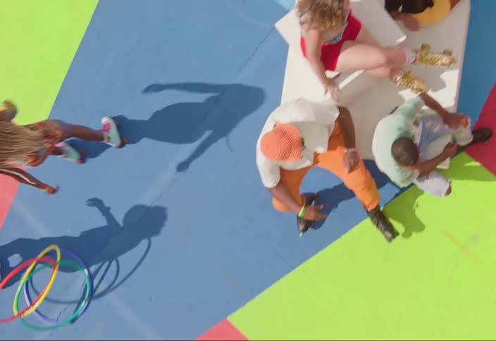 Aerial view of a colorful outdoor scene with people skating and sitting on a surface painted in vibrant pink blue green and yellow sections
