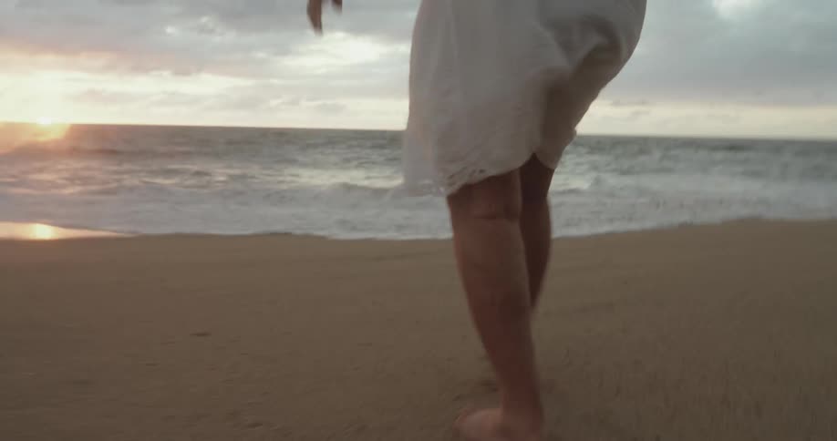 The legs of a person wearing a white dress walking on the sand towards the water of a beach where a wave wets their feet