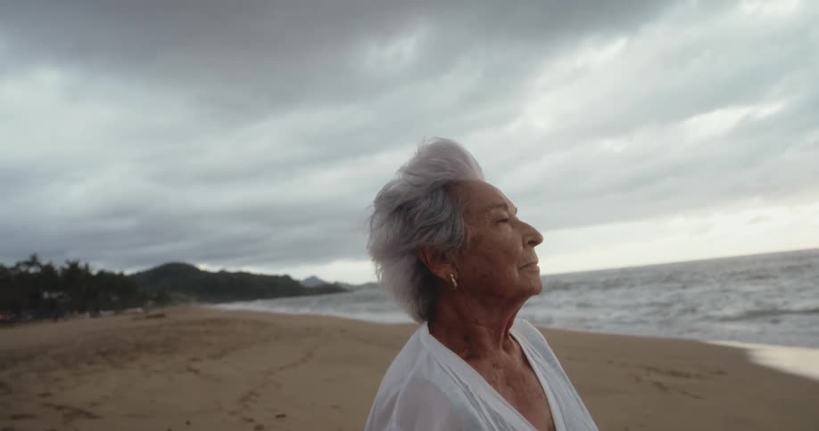 An elderly woman wearing a white outfit stands on a beach staring straight ahead
