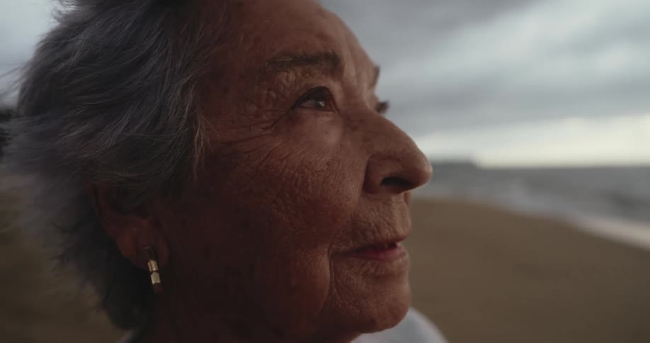 A close up of the face of an elderly woman standing on a beach looking at the sky