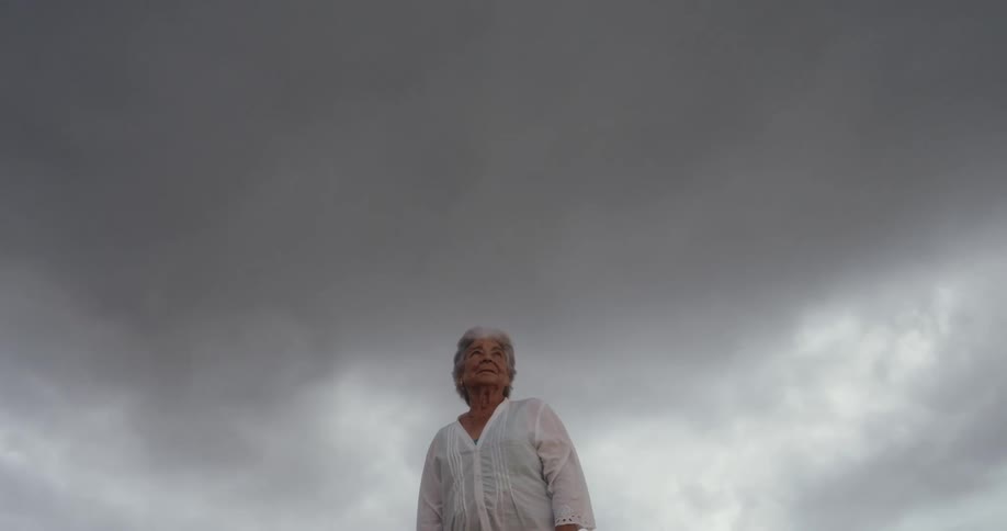 An elderly woman wearing a white blouse stands under a gray overcast sky