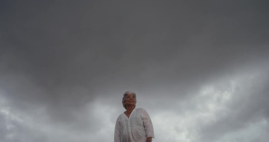 An elderly woman wearing a white blouse stands under a gray overcast sky