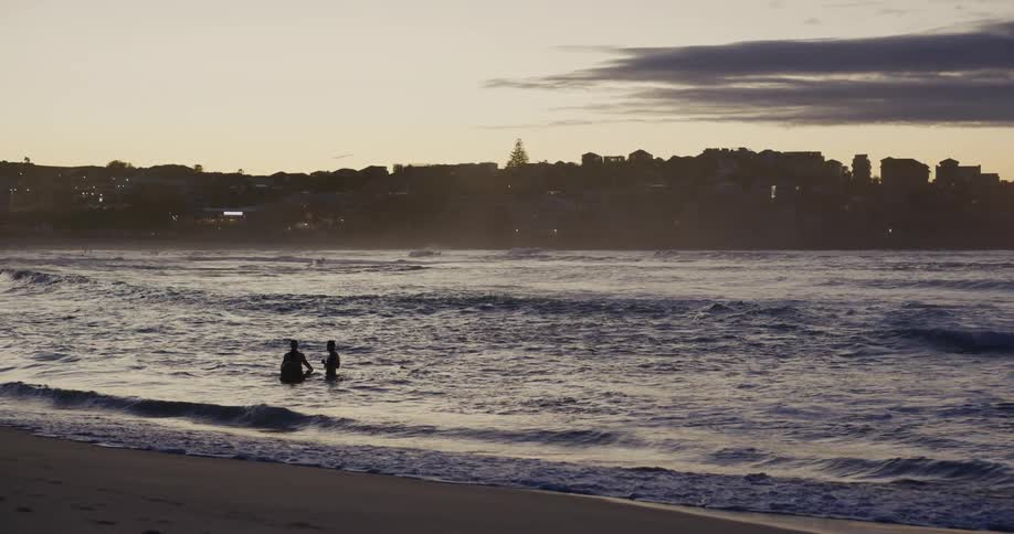Silhouette of two people in the water of a beach with buildings in the background and a late afternoon sky