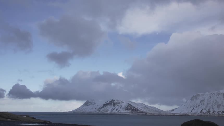 Body of water in the foreground with snowcapped mountains in the background under a cloudy sky