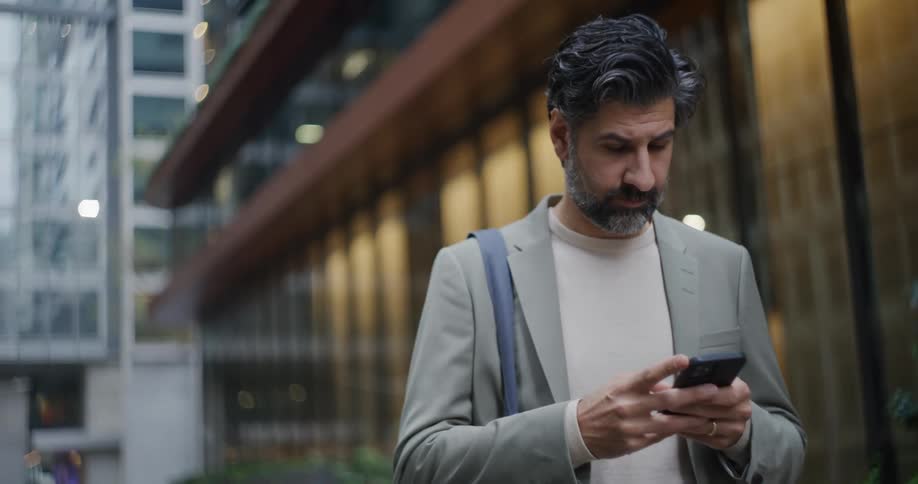 Man standing next to buildings using his smartphone and then putting it inside his gray blazer