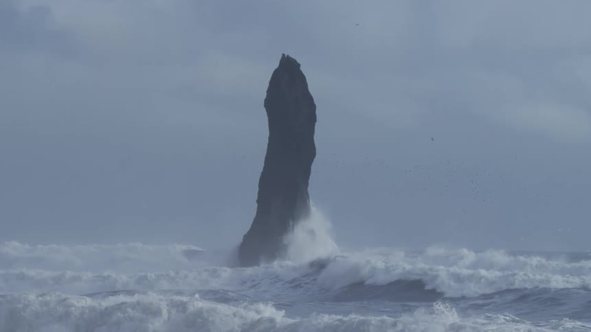 Waves in a stormy sea crash against a towering rock formation on an overcast day