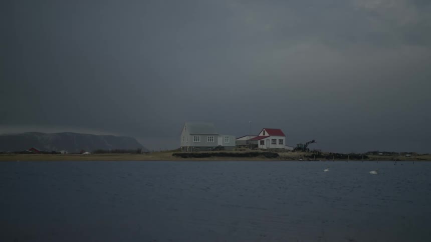 Small houses on the edge of a lake with mountains in the background and a cloudy sky