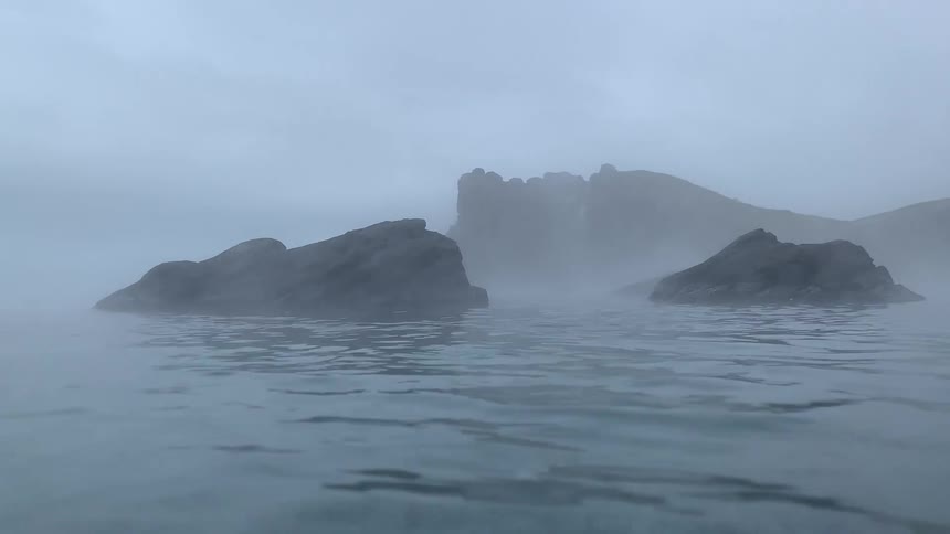 Fog flows over partially submerged rocks near the shore on an overcast day
