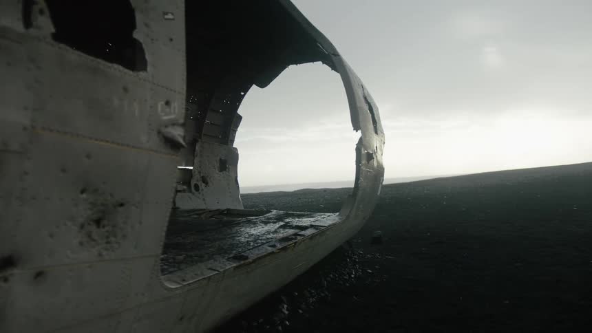 A wrecked airplane fuselage with a gaping hole rests on a dark pebbly ground