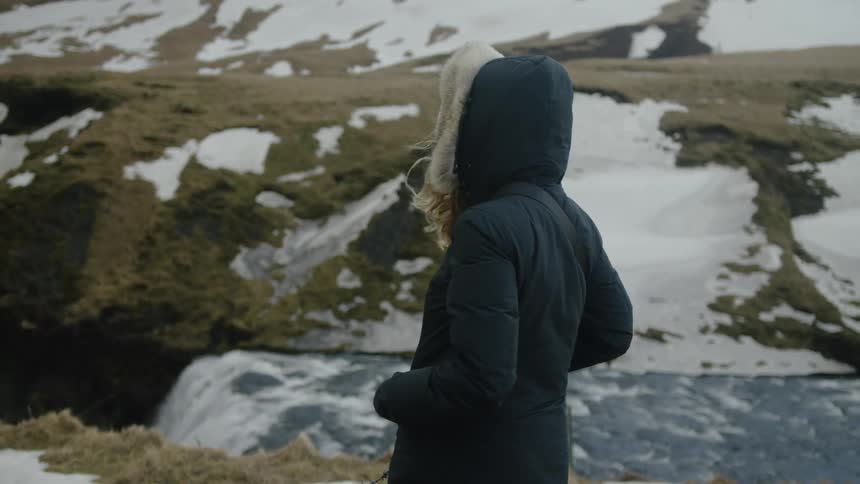 Woman in a hood and jacket and a natural environment with a waterfall next to it on a mountain covered in snow