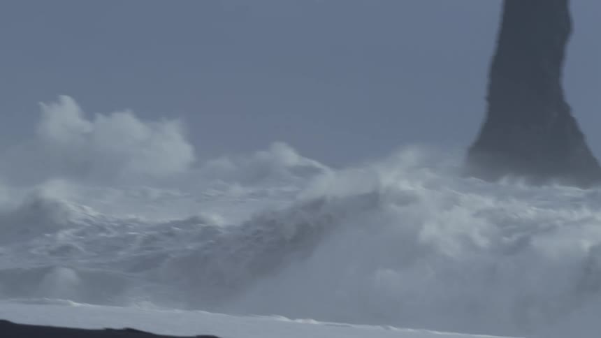 Waves in the ocean with a distinct spray of water rising to its crest against a cloudy sky