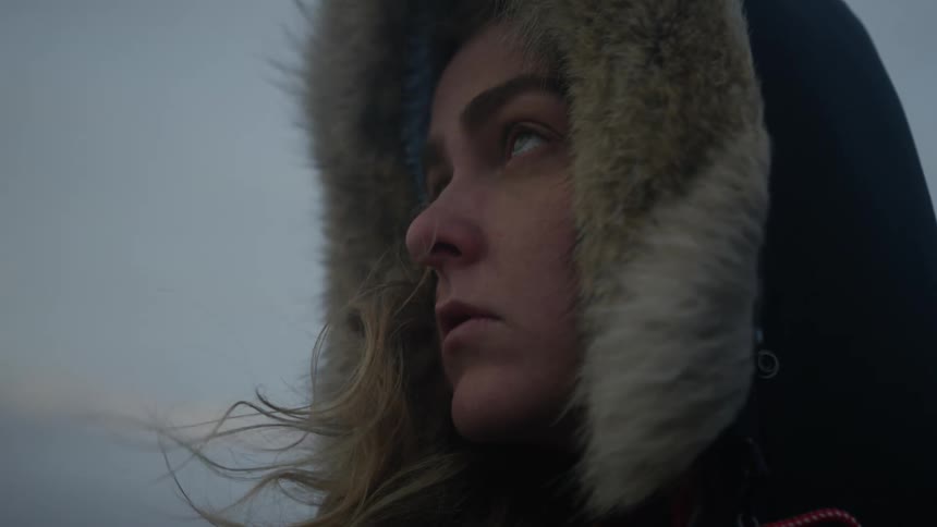 A blonde woman wearing a hood stands outside as the wind blows her hair around