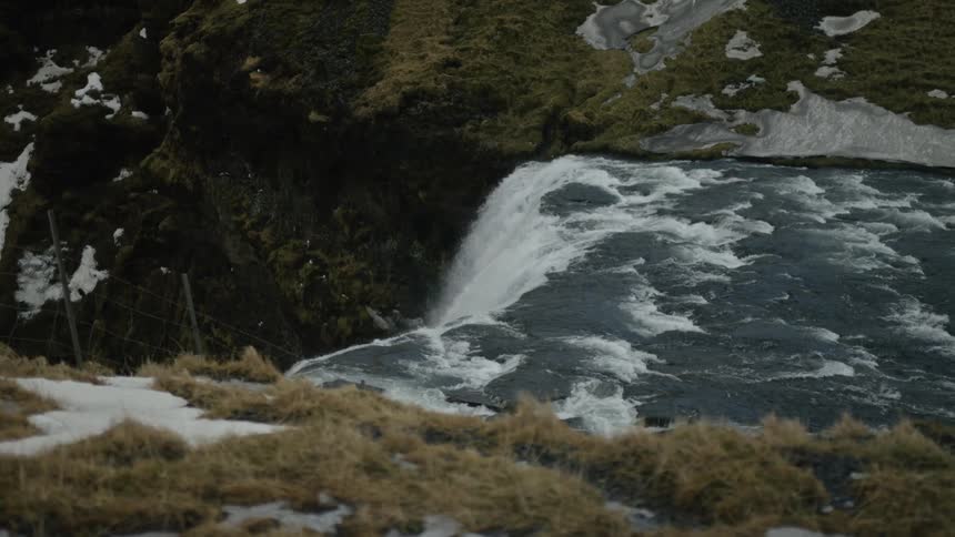 Waterfall flows into a turbulent body of water surrounded by rocky terrain with patches of snow and grass